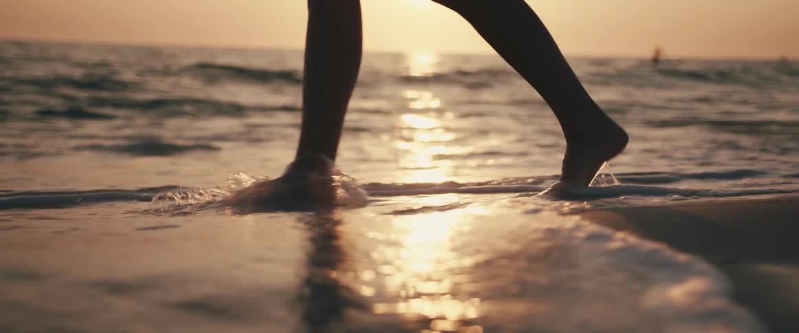 Bare feet walk on wet sand and splash through the surf at sunset