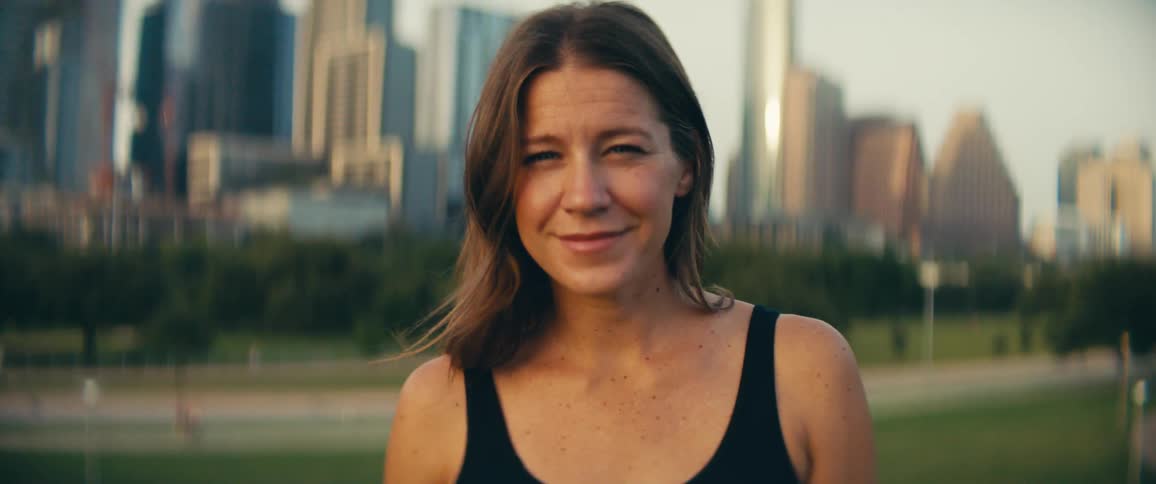 A woman smiles into the camera as the skyscrapers of a metropolis loom behind her on a sunny day