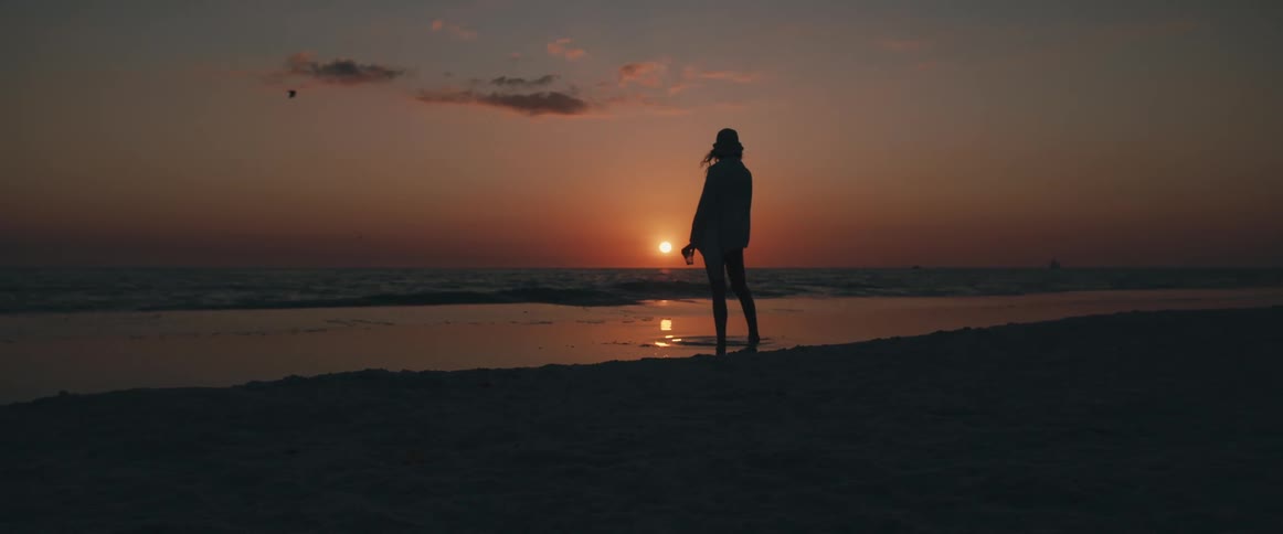 Woman standing on beach next to the sea is silhouetted against sunset