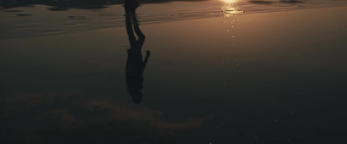 Water pooled on a beach reflects the silhouette of a person walking along the shore at sunset