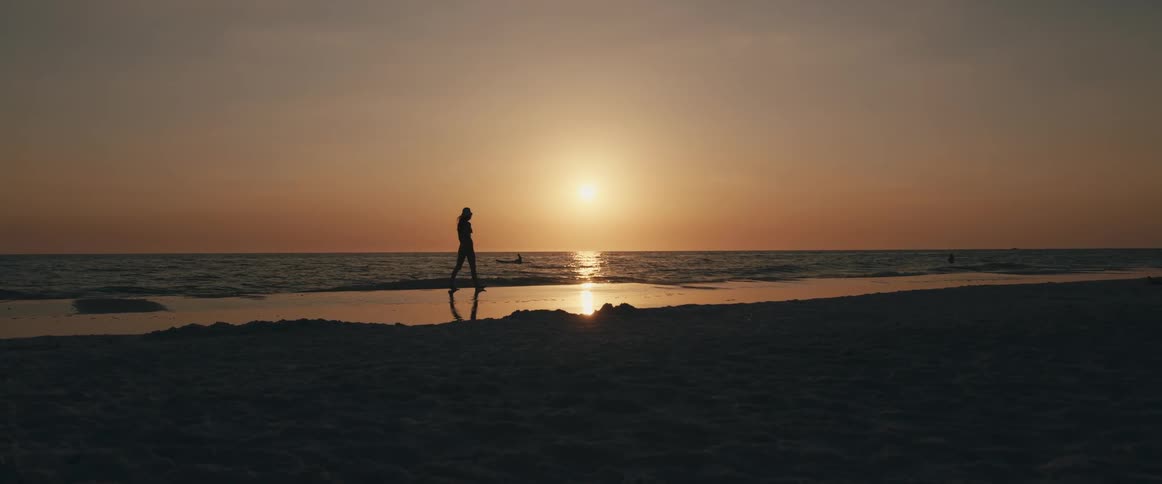 Woman walking on the beach next to the sea is silhouetted against sunset