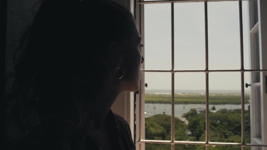 A young woman stands in a dark room and looks out through a barred window at the lake outdoors