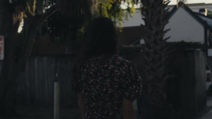 Setting sun shines on person walking down sidewalk past high fence toward a row of houses