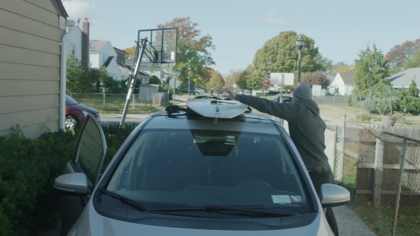 A man moves a strap that's holding a surfboard to the top of a car and walks away