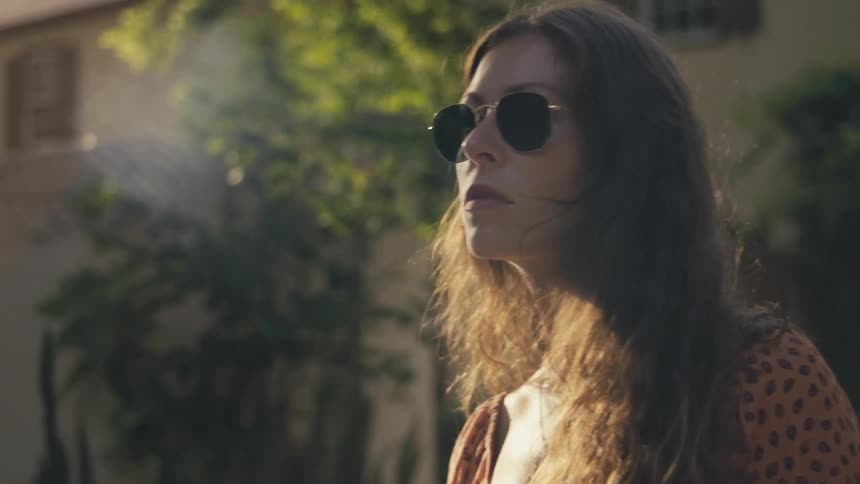 A young woman in fashionable sunglasses standing outside of a house turns to look into the camera