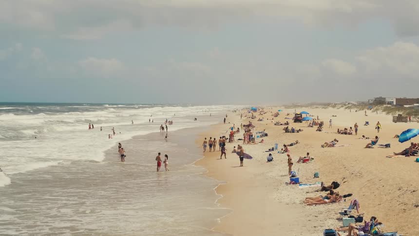 A crowd of people are on a brown sandy beach sunbathing and swim in the ocean