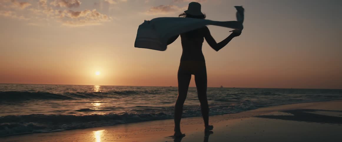 Woman wraps towel around her shoulders while walking along beach looking toward sun sitting on horizon
