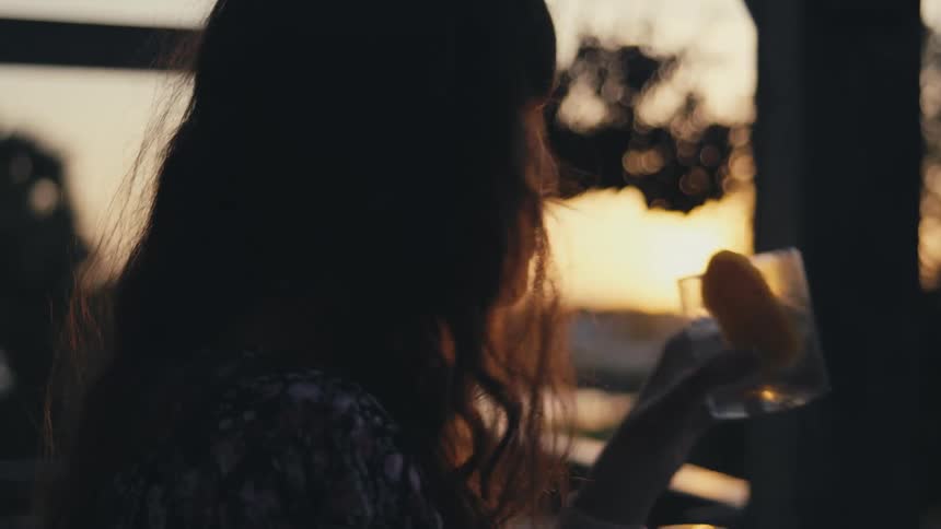 A woman glances at the setting sun while she drinks on her balcony