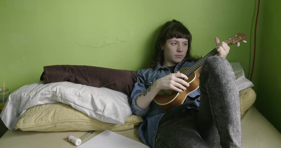 A woman plays a small guitar on the bed