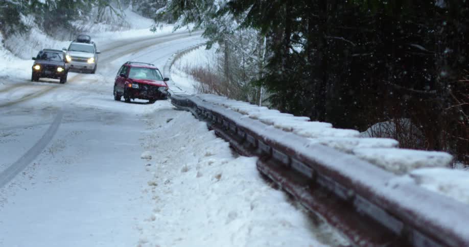 Two cars slowly drive by a smashed up car at the side of the road in a snowbank