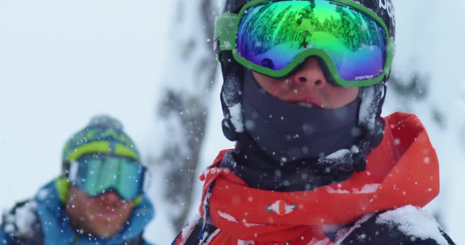 Two men with goggles stand outside in the falling snow