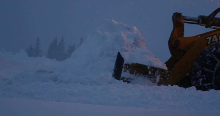 A large snow plow truck plowing snow as it snows outside