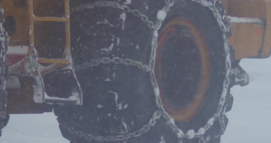 A large vehicle with tire chains drives through a snow storm