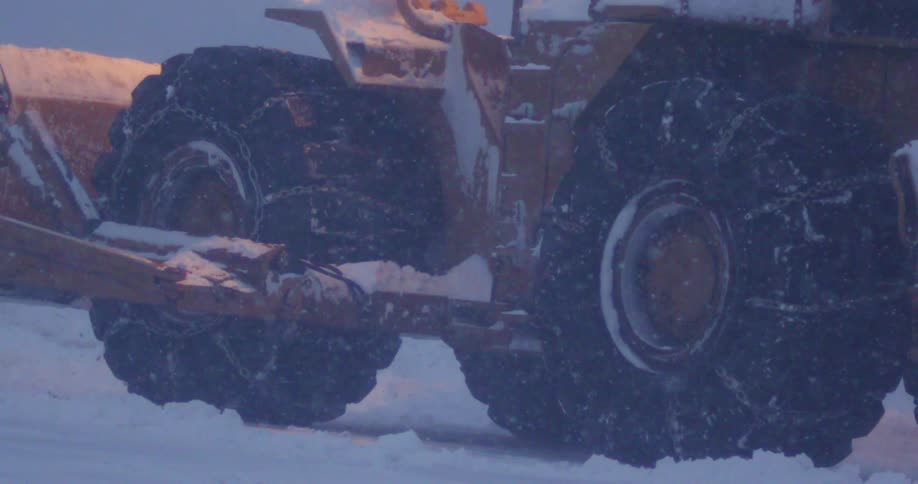 Yellow snow plow with chains on the tires drives backwards n a snow storm
