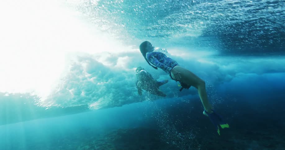 A woman swimming under a wave next to a person holding a camera