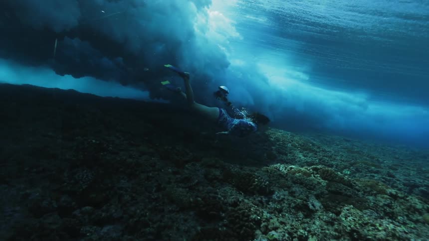 Woman swims underwater near seafloor under shadow of wave passing overhead
