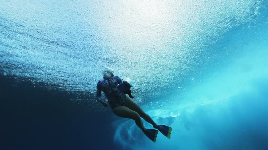 Underwater footage of a diver with a camera surfacing after an ocean wave passes overhead
