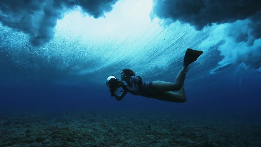 A female diver with an underwater camera films an ocean wave from beneath the sea as it rolls overhead