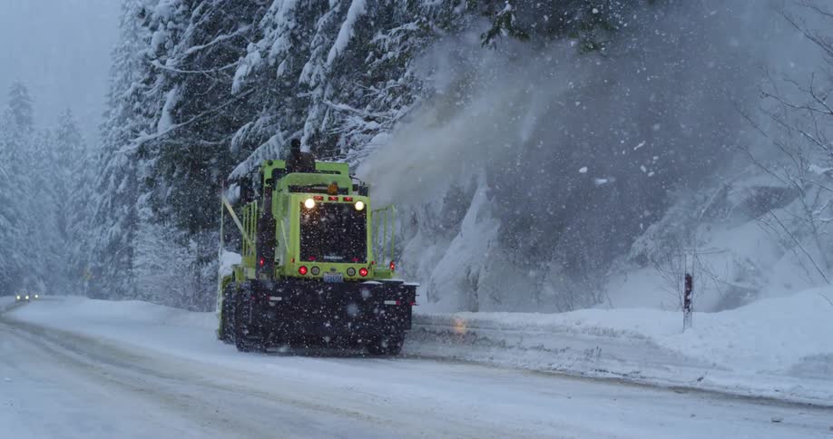 A snow plow clearing a mountain road during a blizzard as a car drives by