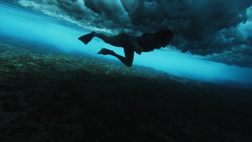 A woman swims over the ocean floor with a huge wave crashing above her