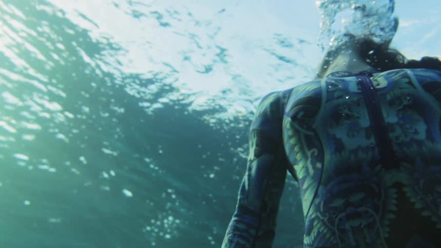 A woman in a patterned wet suit breathes out as she swims towards the surface of the sea
