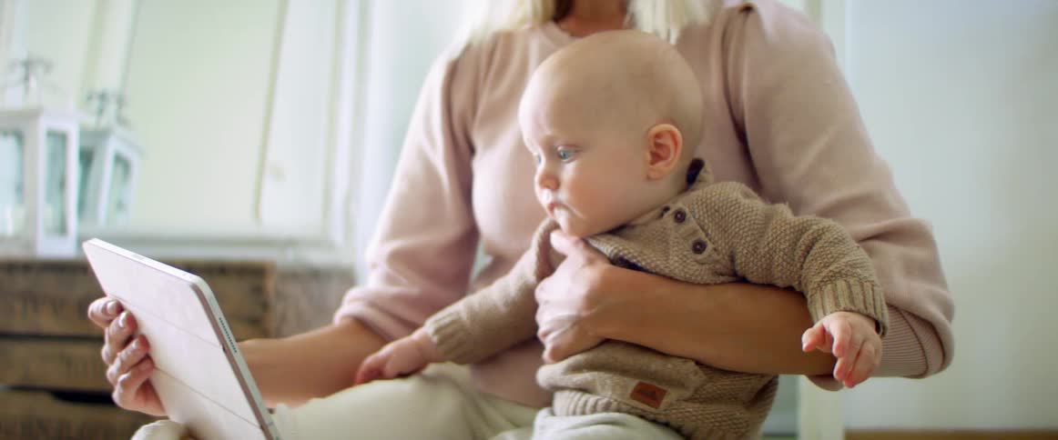 A woman holds an infant on her lap and they watch a tablet screen together