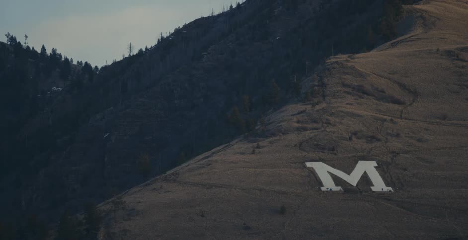 A large letter m sits in the soil on the side of a hill near a forest in Montana