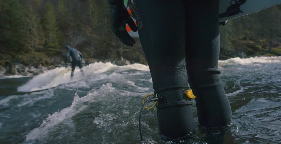 Man wearing black wet suit rides a surfboard on the waves of a river in the mountains