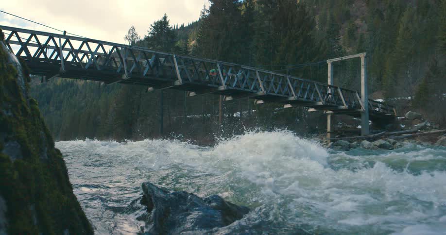 Surfers walk on a narrow bridge over fast moving river water