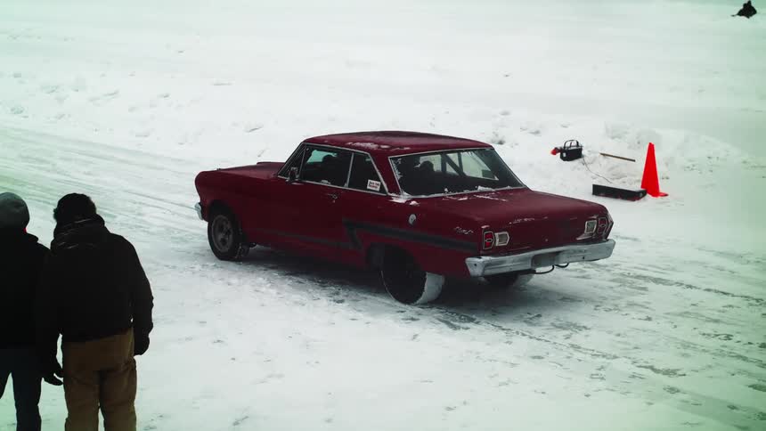 A red car in a snow covered road driving off with two people in the foreground and one person in the background