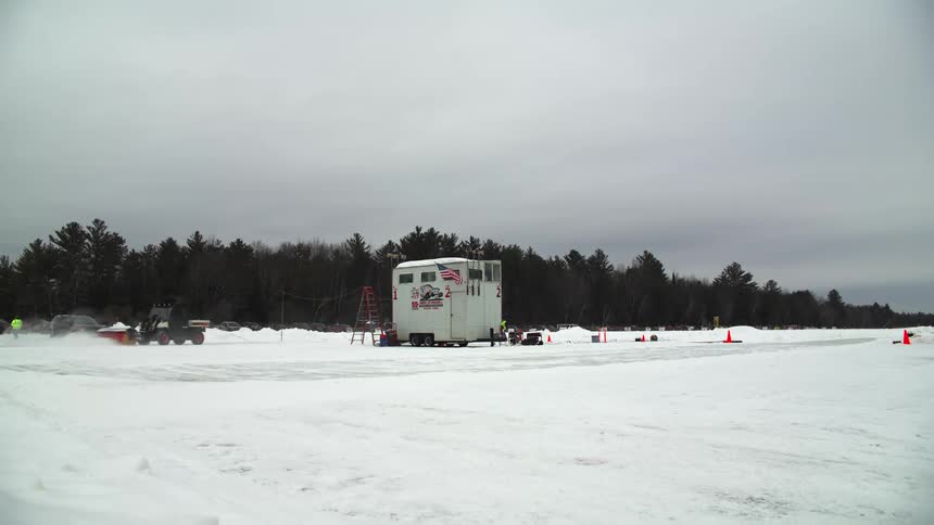 A plow attached to a tractor clears snow from a road next to a white trailer home near a forest