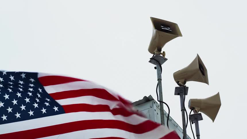 American flag waving in front of speakers atop of poles in front of a building covered in snow
