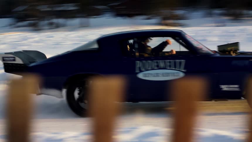 Blue car with company decal drives along snow covered road past person and buildings