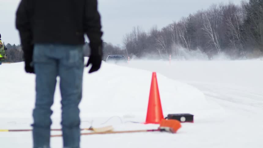 A dark colored older coupe vehicle takes off in the snow down a road marked off with orange cones as people watch