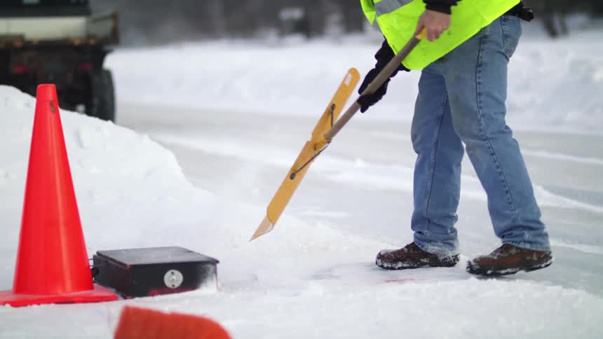A man in a fluorescent vest shovels snow on the side of a cleared road