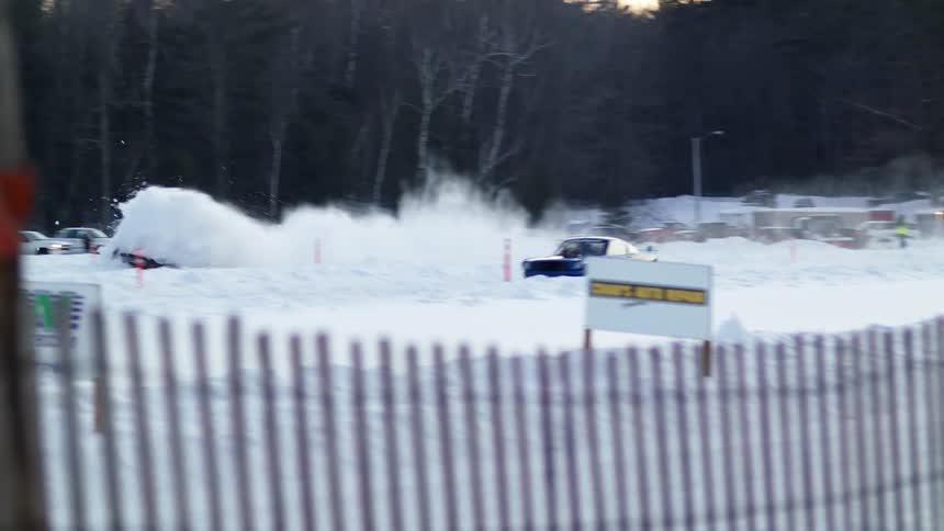 A group of people watch as two cars take off in the snow and race and one car gets stuck in the snow