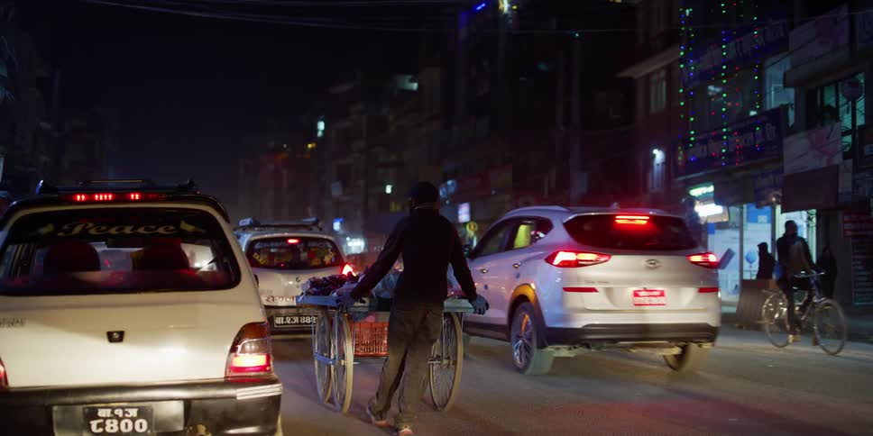 A person pushes a large cart in a lane of heavy city traffic at night