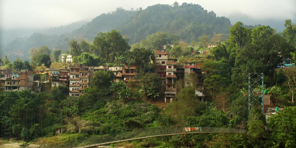 A narrow walking bridge sits in front of a village where apartment buildings and homes are built on the side of a hill near mountains