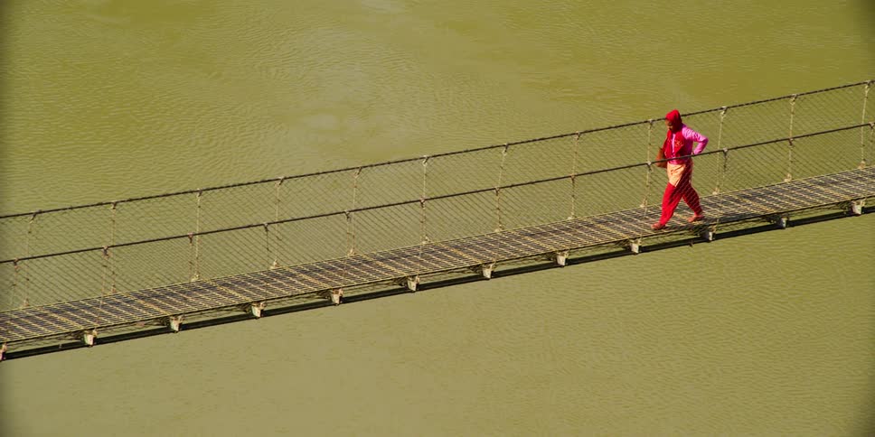 A woman crosses a long narrow bridge walking bridge across a wide brown river during the daytime