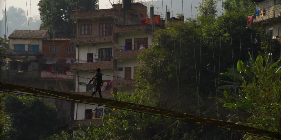 A young woman wearing a backpack walks across a simple suspension bridge to reach a village