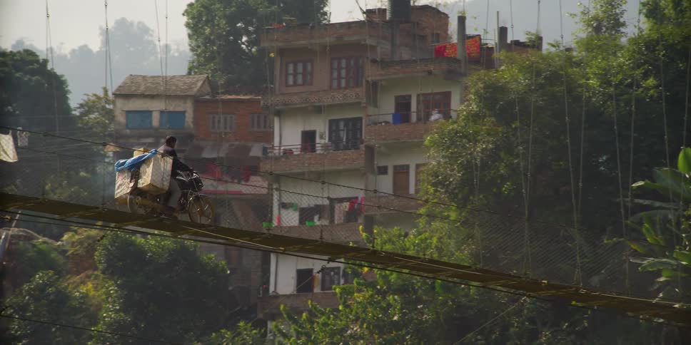 A man with his motorcycle loaded with bags rides across a rickety bridge in a poor neighborhood