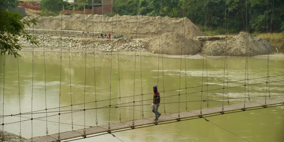 A man walks across a suspension bridge and a group of people sit on the shore