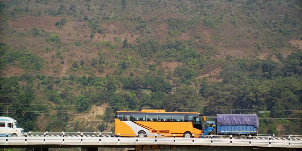 Tour buses delivery trucks and other vehiclesas well as a pedestrian pass each other crossing bridge near vegetation covered slope