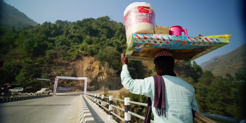 A man walks across a bridge carrying a tray with supplies on his head