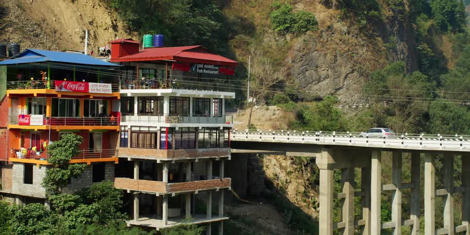 Cars drive over a bridge next to tall buildings half finished on the slopes of a mountain