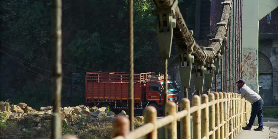 A man with a hat leaning on a handrail on a bridge with a truck and trees in the background