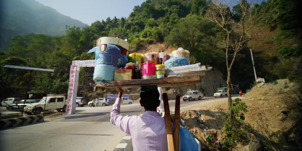 A man is balancing several items on his head while he walks next to a road in the mountains