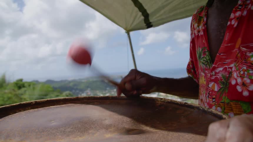 Man plays drums under awning with town spread in valley between hills and water