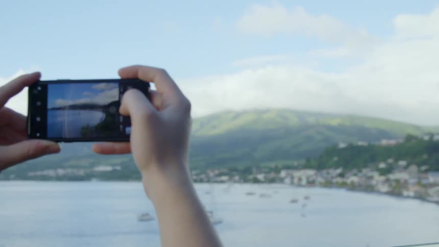 A person standing on a hill uses their smartphone to take a photograph of the bay and city around it below