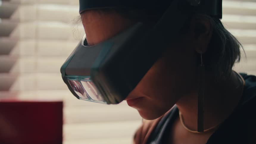 A woman with a magnifying glass and a necklace looking downwards with a window and blinds in the background